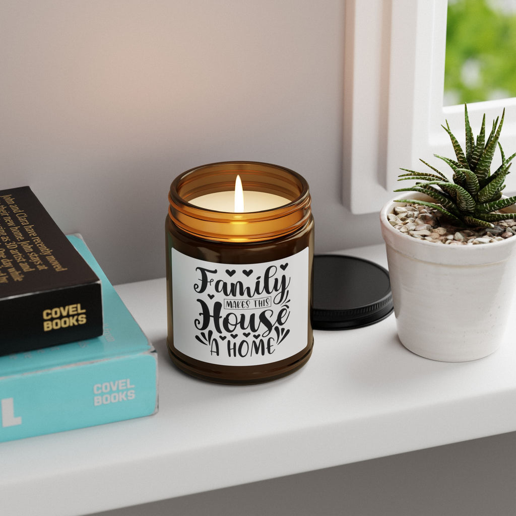 An amber glass jar candle with a white label reading 'Family Makes That House A Home' sits on a white windowsill next to a small potted succulent and stacked books.