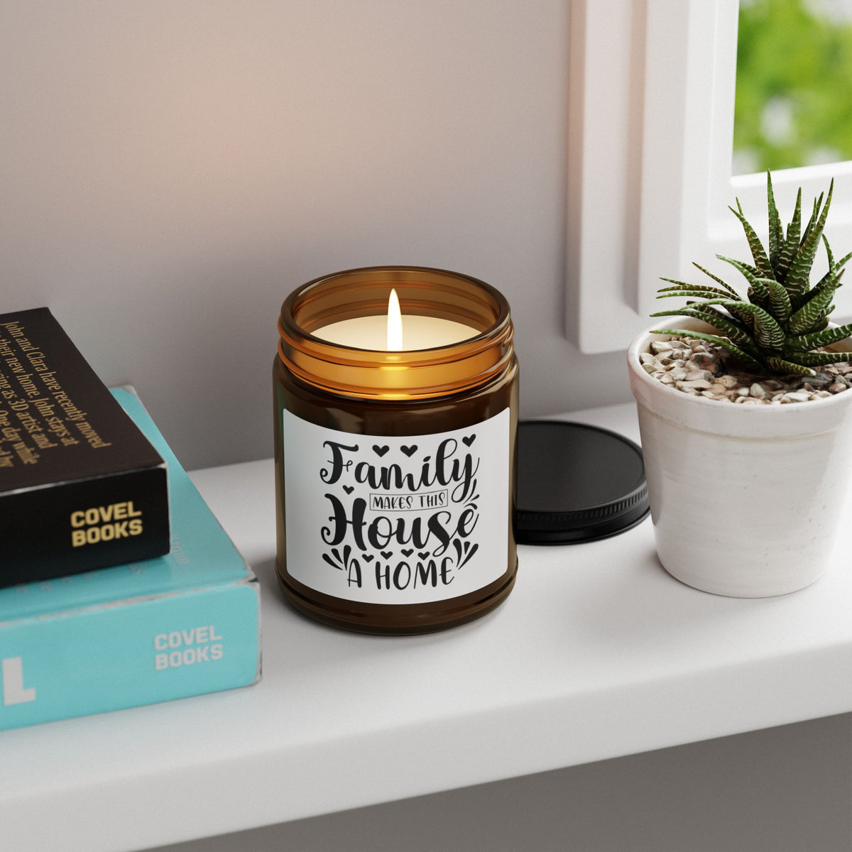 An amber glass jar candle with a white label reading 'Family Makes That House A Home' sits on a white windowsill next to a small potted succulent and stacked books.