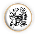 A round wall clock with a light natural wood frame and a white face, featuring a bold black graphic of a skull and skeletal hand making a middle finger gesture, with the text 'LIFE'S TOO SHORT TO GIVE A SHIT,' on a clean white background.