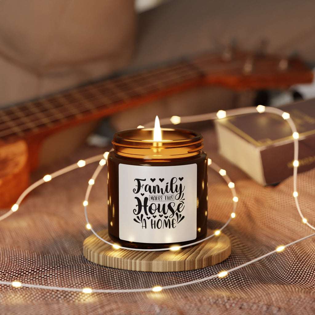 A lit amber glass jar candle sits on a small wooden coaster, wrapped with warm white fairy lights in a cozy indoor setting. The candle has a white label with black text reading 'Family Makes Up House of Home.