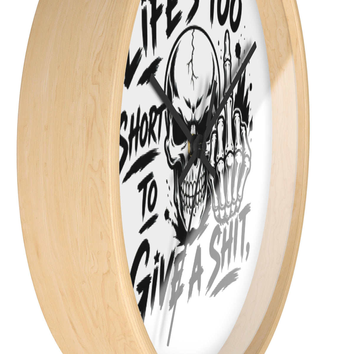 A round wall clock with a light natural wood frame and a white face featuring a black graphic of a skull and skeletal hand making the middle finger, alongside bold text reading 'LIFE IS TOO SHORT TO GIVE A SHIT'.