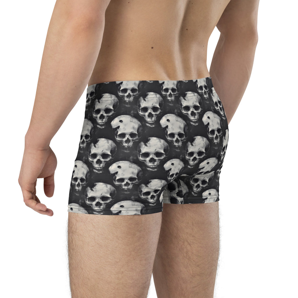 Rear view of a male model wearing boxer briefs with an all-over grayscale skull print on a dark background, against a plain white studio backdrop.