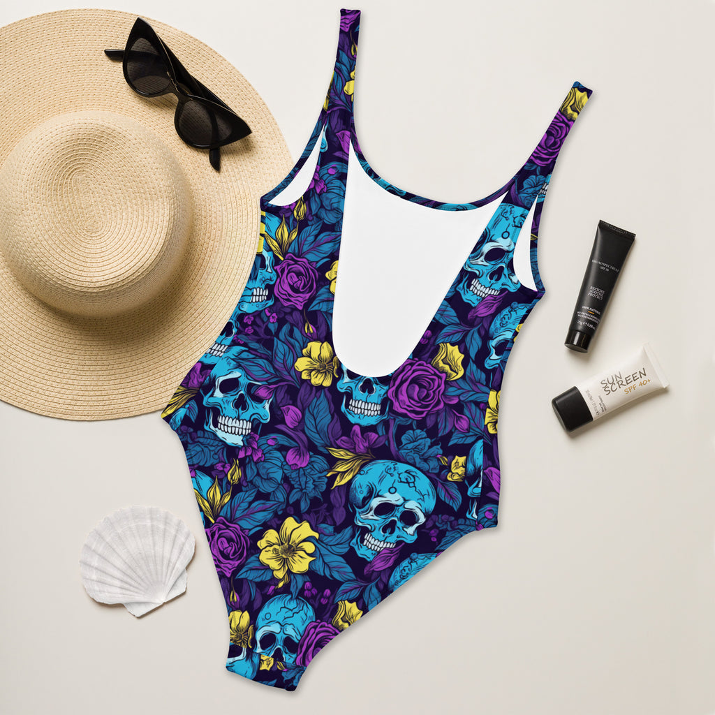 A one-piece swimsuit with a vibrant blue and purple floral and skull pattern, styled in a flat lay with a straw hat, black sunglasses, a white seashell, and two tubes of sunscreen on a light beige background.