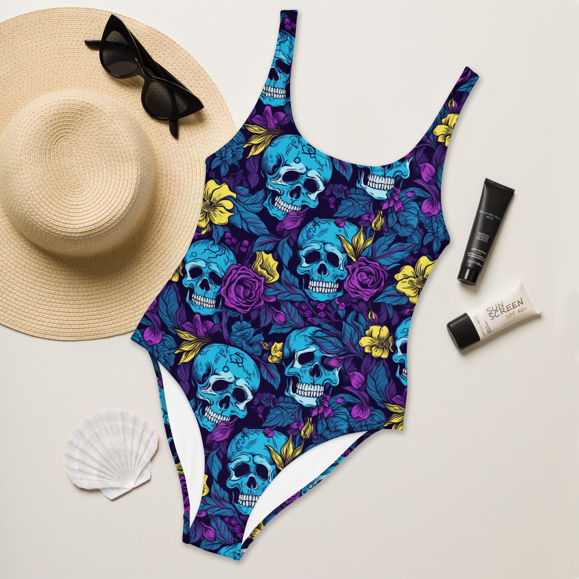 A one-piece swimsuit with a vibrant blue and purple skull and floral print, styled in a flat lay with a straw hat, black sunglasses, a white seashell, and two tubes of sunscreen on a light beige background.
