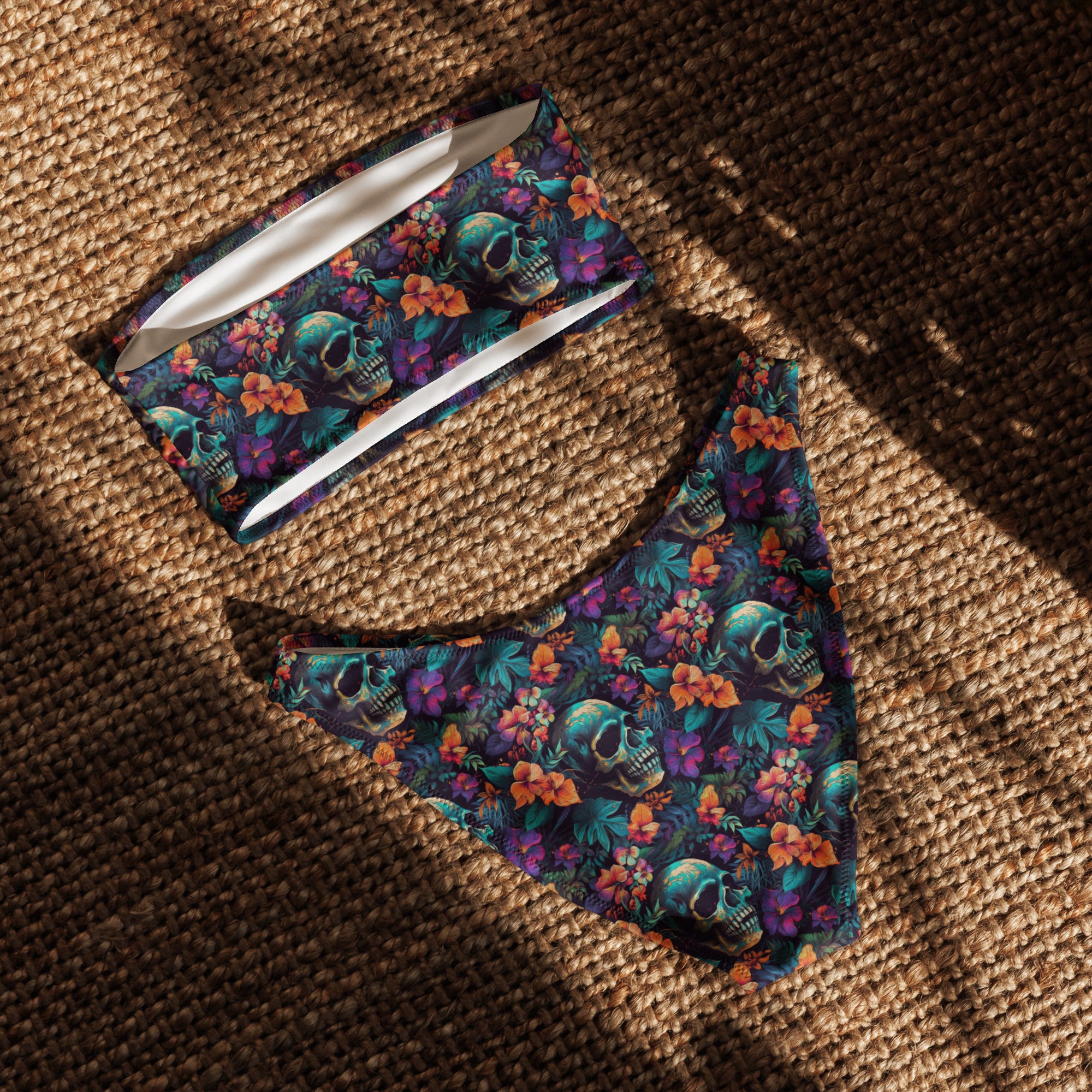 A two-piece bikini set laid flat on a brown woven rug under dappled sunlight. The set features a dark teal background with a vibrant tropical floral and skull print in purple, orange, and green.