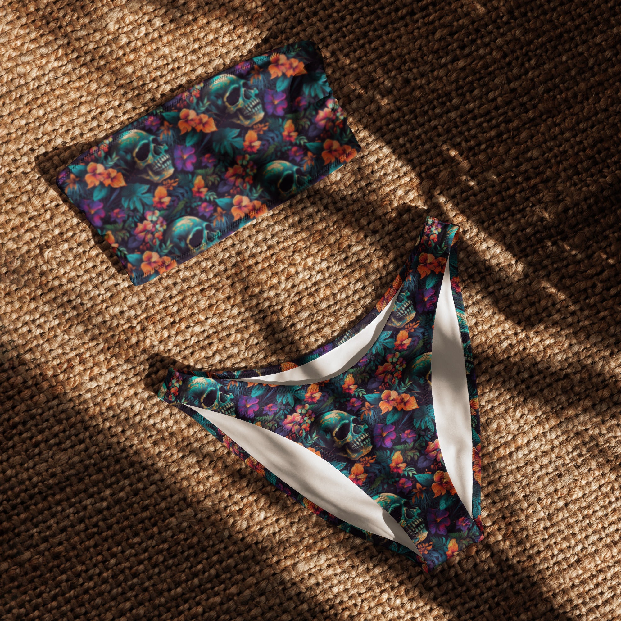 A two-piece bikini set laid flat on a woven straw surface under dappled sunlight. The set features a dark tropical print with green skulls, orange flowers, and teal foliage, with contrasting white side panels.