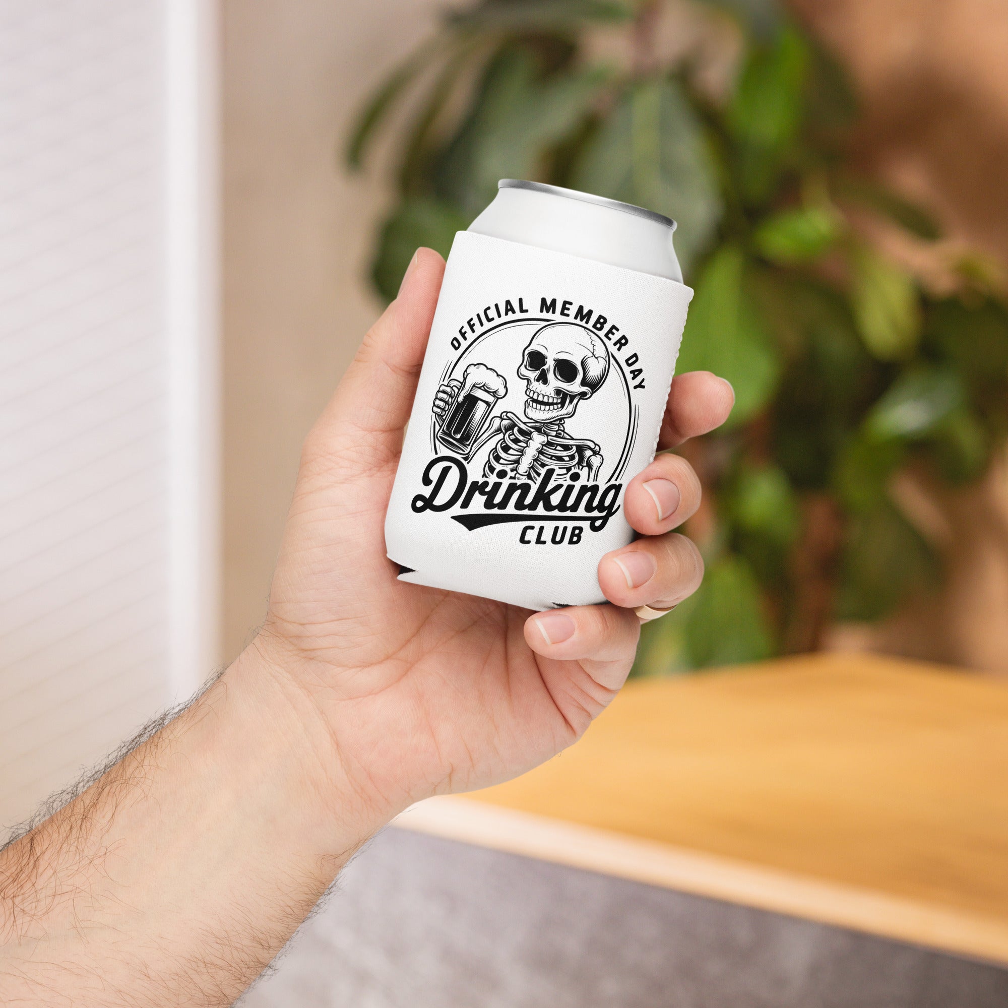 Hand holding a can cooler with a skeleton design and text, blurred indoor background