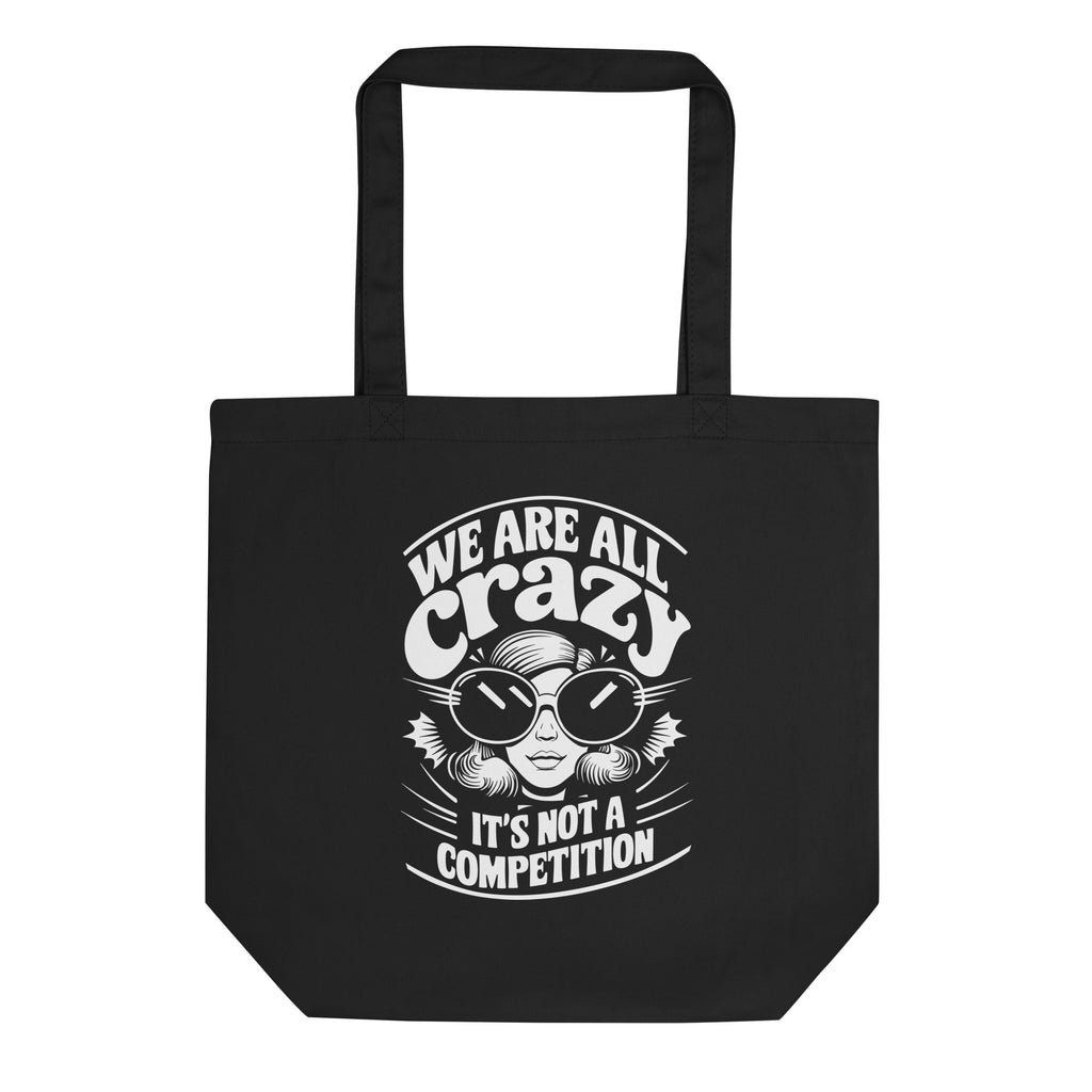 A black canvas tote bag with black handles, featuring a white graphic print of a cartoon character wearing sunglasses and the text 'WE ARE ALL crazy IT'S NOT A COMPETITION'.