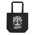 A black canvas tote bag with black handles, featuring a white graphic print of a cartoon character wearing sunglasses and the text 'WE ARE ALL crazy IT'S NOT A COMPETITION'.