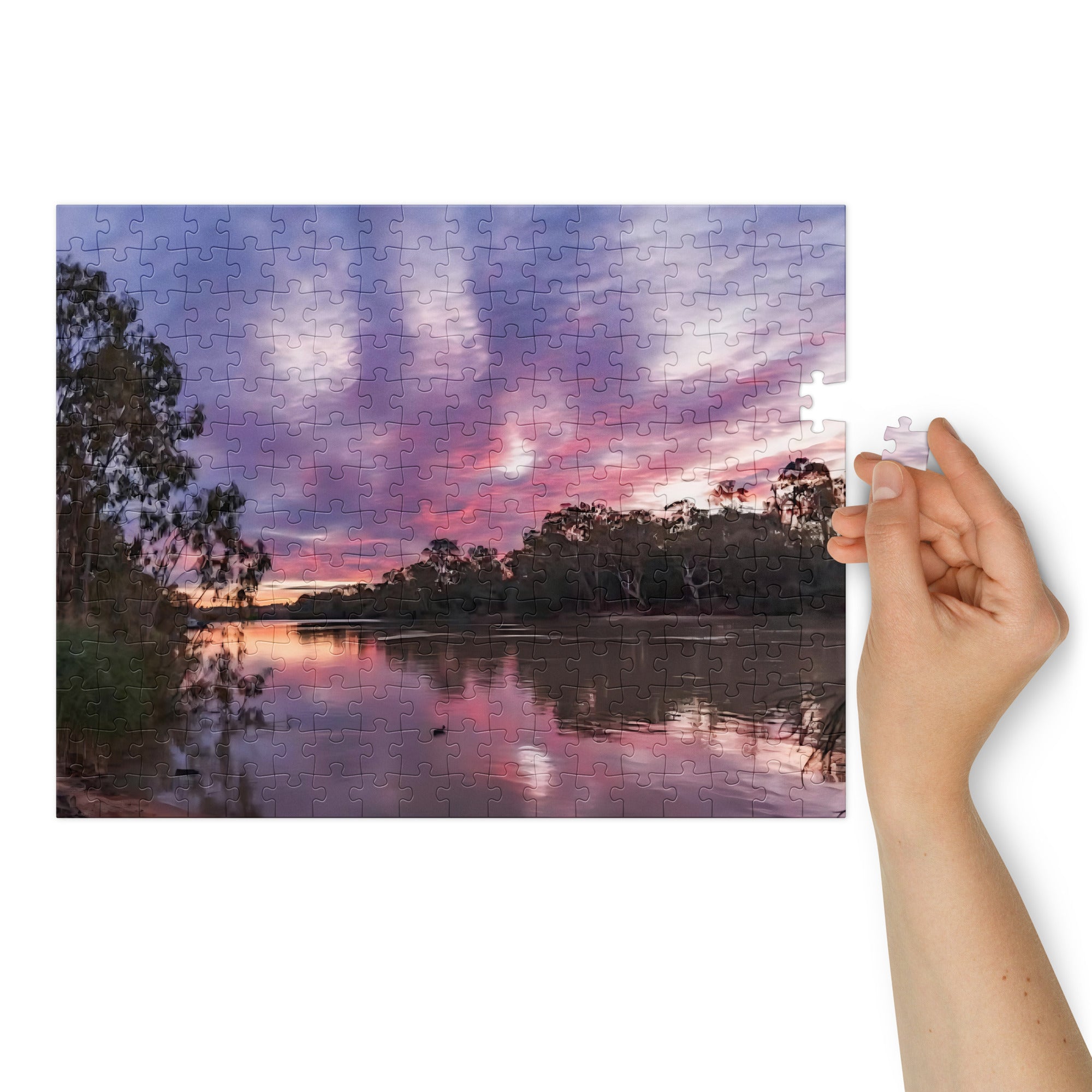 Jigsaw puzzle with a sunset over a lake and trees, held together by a hand.