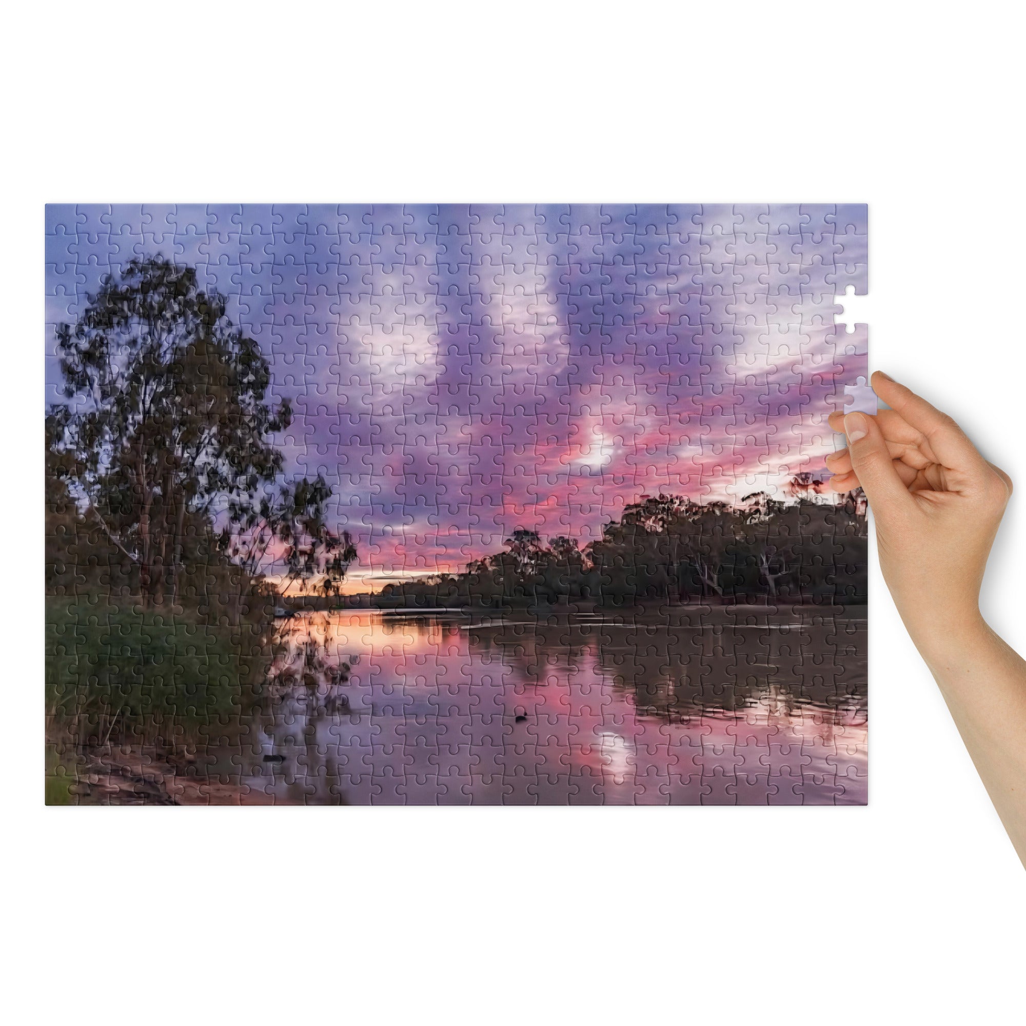 Jigsaw puzzle piece with a sunset over a lake and trees, held by a hand.