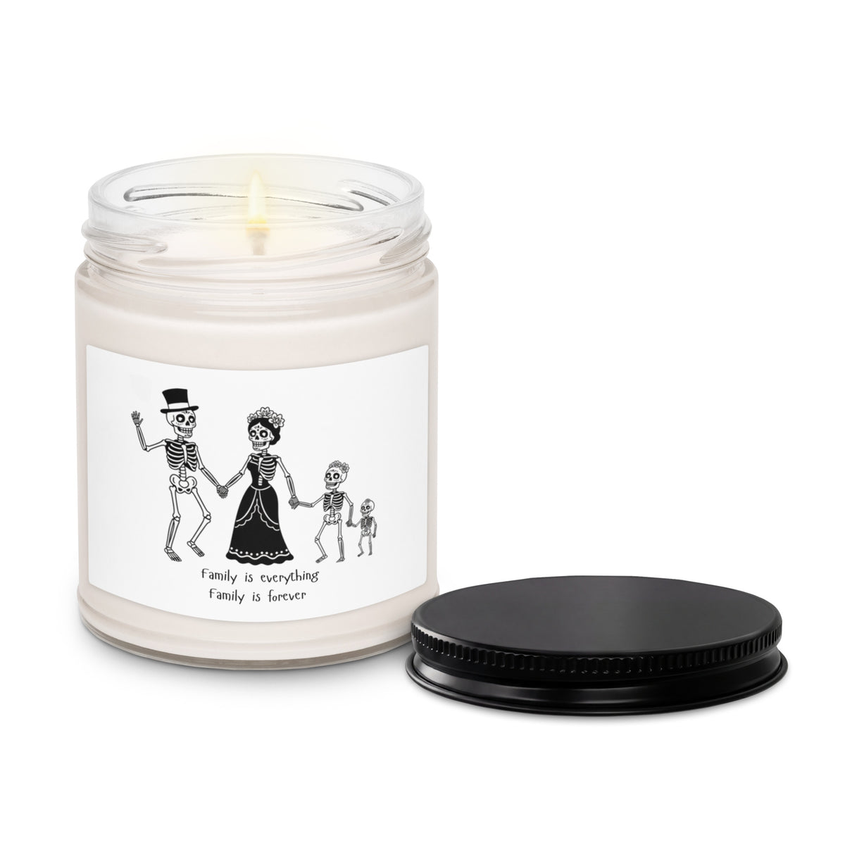A white wax candle in a clear glass jar with a lit wick, featuring a black-and-white illustration of skeleton family members on the label. A black screw-on lid sits beside the jar on a clean white background.