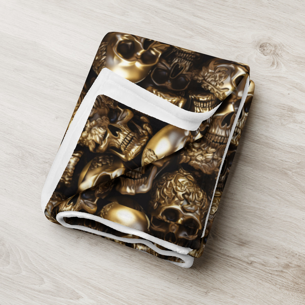 A folded blanket featuring a metallic gold skull and crossbones pattern on a black background, with a white plush trim, displayed on a light wood floor.