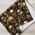A folded blanket or throw featuring a repeating metallic gold skull pattern on a black background, with a white reverse side, displayed on a light wood floor.